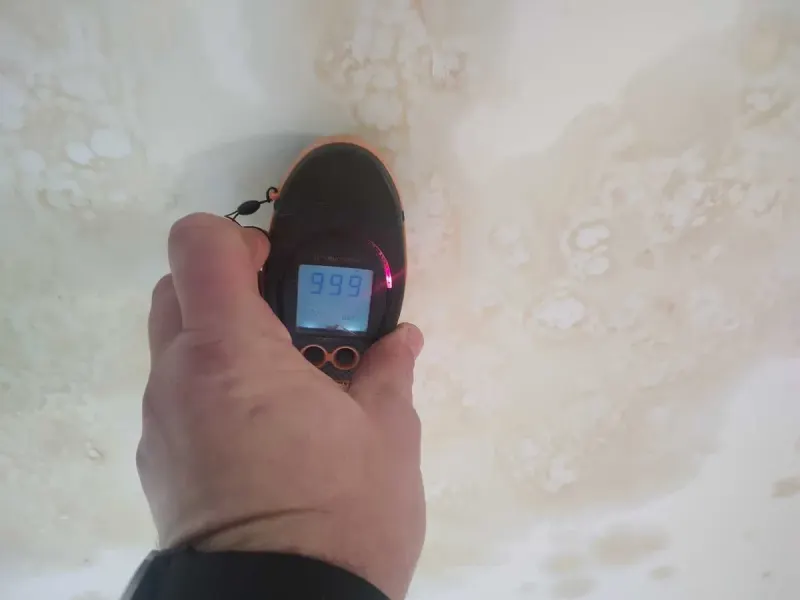 A damp meter held against the stained living room ceiling
